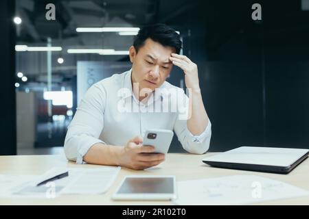 Worried young Asian man sitting in office at desk and using mobile phone. Upset, he looks at the screen, holds his head. Stock Photo