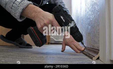 fixing a dark wooden floor plinth with a screwdriver in a man's hand, fixing a brown plinth between a floor with gray linoleum and the wall Stock Photo