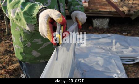 a man in gloves cuts a polyethylene covering material with a clerical knife on the street, cutting a polyethylene dense film with a knife Stock Photo