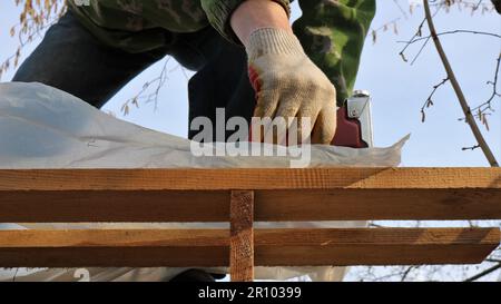 fixing plastic film on the wooden floors of the roof of a barn frame using a construction stapler, a construction gun in the gloved hands of a builder Stock Photo