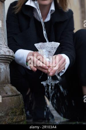 Woman in office suite holds with two hands Martini cup. water falls in a crystal glass. Girl fills a glass with water from a street fountain. Drinking Stock Photo
