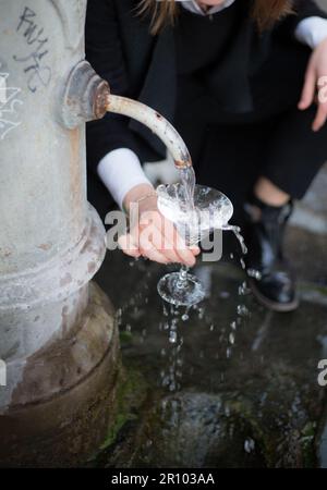 Traditional roman drinking fountain. Woman holds Martini cup. water falls in a crystal glass. Background blurred. Girl fills a glass with water Stock Photo