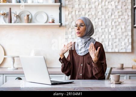 Very hot at home. broken air conditioner. Young woman in hijab sitting at home in front of laptop and waving hands at face, making fresh cool air, suffering from heat, breathing hard, panic attack. Stock Photo