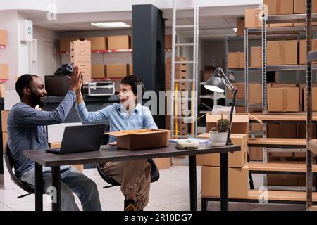 Happy employees doing high five in storehouse after finishing packing clothes orders in cardboard box, celebrating triumph. Smiling diverse team discussing transportation logistics in warehouse Stock Photo