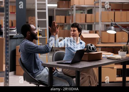 Happy workers doing high five in storehouse after finishing packing helmet orders in cardboard box, celebrating triumph. Smiling diverse team discussing transportation logistics in warehouse Stock Photo
