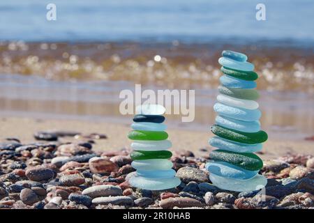 Two balanced pyramids of glass bottle fragments polished by the sea against the background of the oncoming wave. Summer vacation by the sea Stock Photo