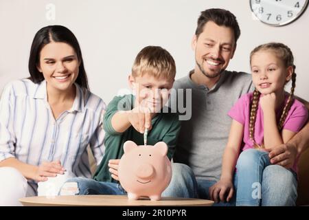 Happy family putting money into piggy bank at table in room Stock Photo