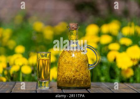 Homemade tincture of dandelion flowers in a shot glass and in a glass bottle on a wooden table in a summer garden, close up Stock Photo