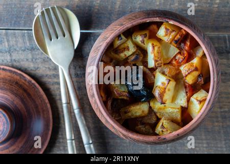 Stewed potato, carrot, onion, tomato and prunes in a clay pot on wooden table background, close up, top view Stock Photo