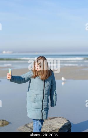Smiling young woman in blue parka walking near sea at sunny spring day. Taking selfie Stock Photo