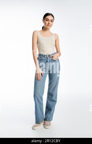 full length of charming young brunette woman standing in blue denim jeans and tank top while posing with hand in pocket and looking at camera on white Stock Photo