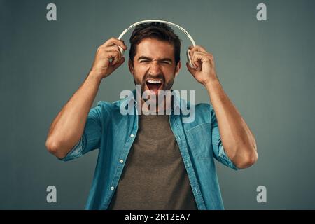 Blasting some beats. Studio shot of a handsome young man screaming while holding headphones against a grey background. Stock Photo