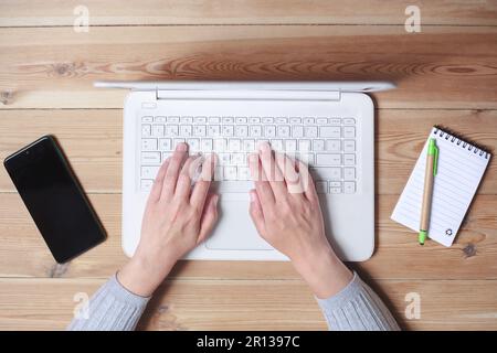 Top view of a woman's hands typing on a laptop. Next to them is her smart phone and a notepad. Stock Photo