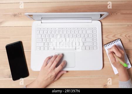 Woman's hands are visible on a laptop keyboard and writing in notebook. Stock Photo