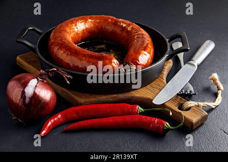 Grilled sausage rolled into a ring on a cast-iron pan with cutlery, a wooden cutting board, hot peppers and red onions on a black stone background Stock Photo
