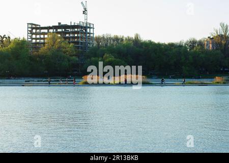 Сonstruction of a multi-storey building in park area, where people walk, there is a lake and green trees. Landscape. Focus on center photo Stock Photo