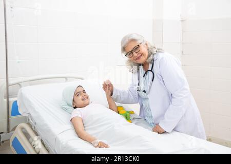 Smiling indian senior woman doctor holding hand of little girl cancer patient lying on hospital bed undergoing course of chemotherapy. Looking at came Stock Photo