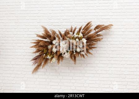A beautiful floral set of white flowers, reeds and feathers in white and brown colors hangs on a white brick wall. And beautifully adorns interior of Stock Photo
