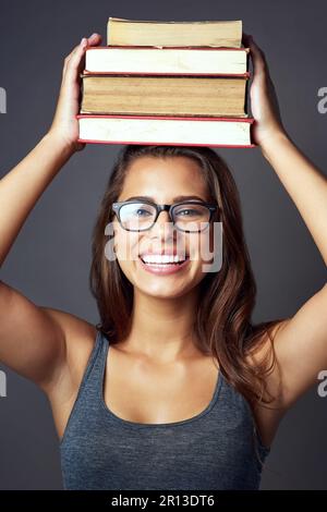 Get your reading on. Studio portrait of a young woman balancing a pile of books on her head against a grey background. Stock Photo