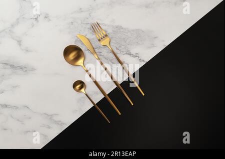 Top view of gold cutlery on white marble and black background. Creative table setting flat lay. Fork, knife, spoon and dessert spoon. Copy space. Stock Photo