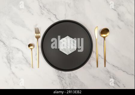 Top view of black plate and gold cutlery on white marble background. Table setting flat lay. Fork, knife, spoon, dessert spoon and plate. Copy space. Stock Photo