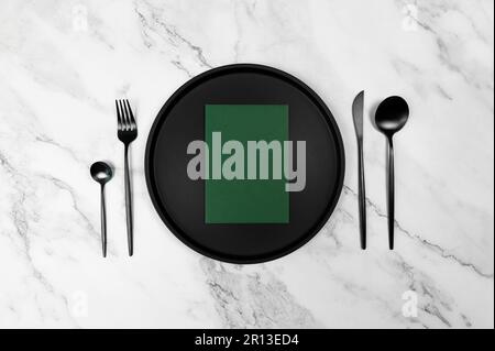 Top view of black plate and black cutlery on white marble background. Table setting menu, empty green paper blank flat lay, copy space. Stock Photo
