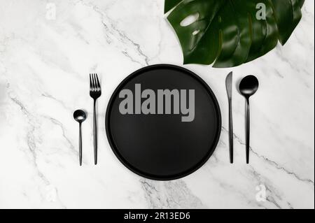 Top view of black plate and black cutlery on white marble background. Luxury table setting flat lay, copy space. Tropical monstera leaf. Stock Photo