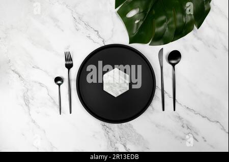 Top view of black plate and black cutlery on white marble background. Luxury table setting flat lay, copy space. Tropical monstera leaf. Stock Photo
