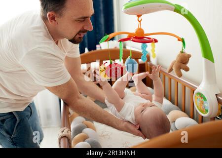 A happy dad puts his baby in a crib with a musical toy. The father hugs his baby. Happy childhood Stock Photo