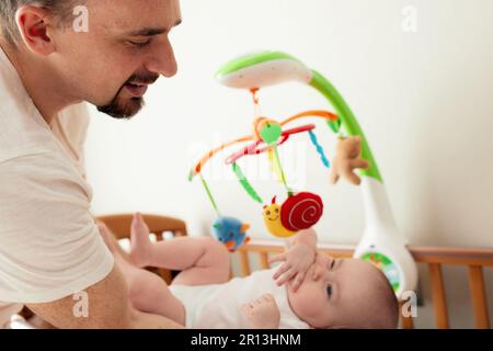 A happy dad puts his baby in a crib with a musical toy. The father hugs his baby. Happy childhood Stock Photo