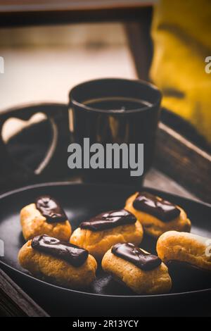 Mini eclairs with dark chocolate and a coffee on a wooden tray, french pastry specialty with custard cream, vertical Stock Photo
