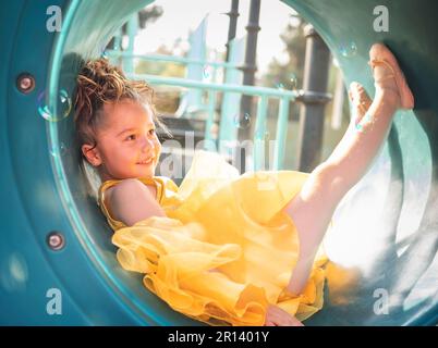 beautiful junior little girl playing in the park in yellow dress laughing with bubbles, screaming with happiness in family enjoying children's day Stock Photo