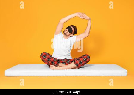 Man in sleeping mask sitting on soft mattress and stretching against orange background Stock Photo