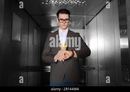 Young businessman with cup of hot drink checking time in elevator Stock Photo