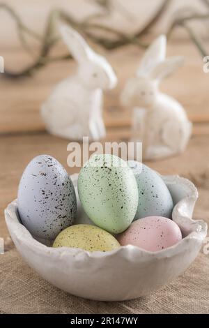 Decorative Easter eggs in pastel colors in a white handmade bowl on a wooden table, two white wooden Easter bunnies in the background Stock Photo