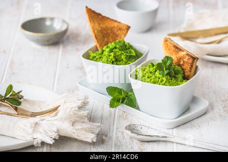 Two bowls of pea puree with basil, mint and roasted bread on white wooden background Stock Photo