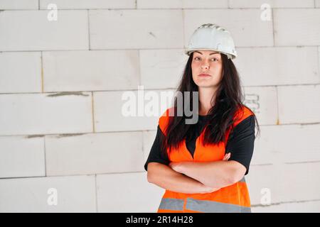 Young woman in white hard hat and orange high visibility vest, long dark hair, hands crossed, looking into camera. Blurred construction site wall back Stock Photo