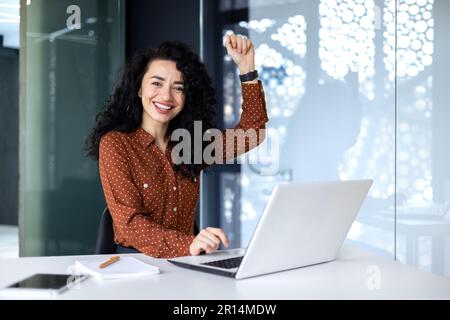 Portrait successful Hispanic business woman office worker with curly hair smiling and looking at camera working at table with laptop holding hand up gesture success and triumph celebrating victory Stock Photo