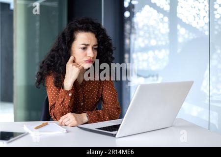 Serious thinking Hispanic businesswoman working inside modern office, female programmer looking at laptop testing new software. Stock Photo