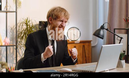 Concentrated young businessman working on laptop at home office desk typing browsing say Wow yes found out great big lottery jackpot win, good news, celebrating. Bearded manager freelancer man job Stock Photo