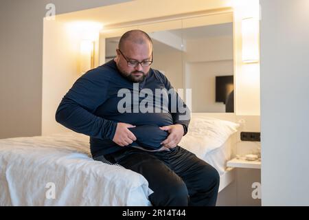 Man suffering from extra weight touching abdomen sitting on bed in bedroom.  Stock Photo