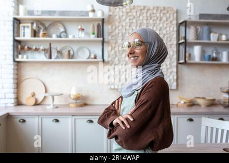 Portrait of a young woman of oriental origin in a hijab and an apron standing in the kitchen at home with her arms crossed on her chest and looking confidently to the side. Stock Photo