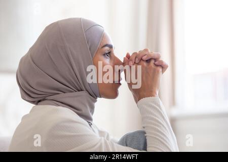 Close-up photo. Portrait of young Muslim woman in hijab looking serious and sad. He sits thoughtfully at home, folding his hands together in front of him. Stock Photo