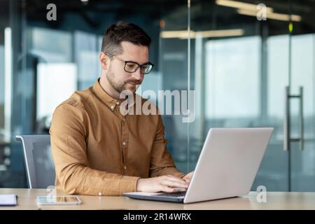Portrait of a young male programmer, in a brown shirt and glasses who is concentrating on working on a laptop, while sitting at a desk in the office. Stock Photo