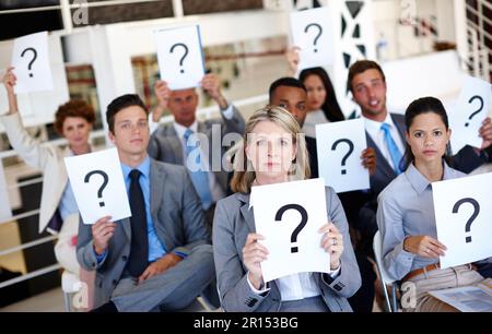 There are so many questions. a group of businesspeople holding up signs with question marks on them during a work presentation. Stock Photo
