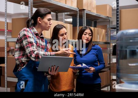 Warehouse employees team cooperating and preparing order using pick ticket on laptop. Storehouse asian workers doing inventory management and solving delivery problems together Stock Photo