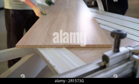 A worker in a furniture factory holds a table top. Furniture maker. A worker stands by a factory machine with a wooden piece while processing Stock Photo