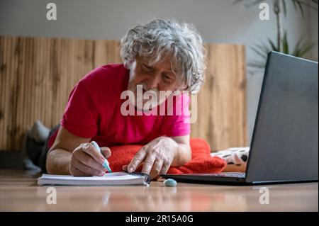 Senior man lying on living room floor taking notes in his notebook as he attends an online class using laptop computer. Stock Photo