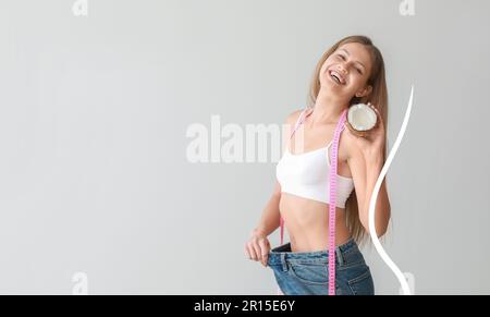 Beautiful young woman in loose jeans, with coconut and measuring tape on light background. Diet concept Stock Photo