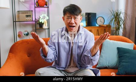 What. Why. Asian man raising hands in indignant expression asking reason of failure demonstrating disbelief irritation by troubles at modern home apartment. Chinese guy in living room sitting on sofa Stock Photo